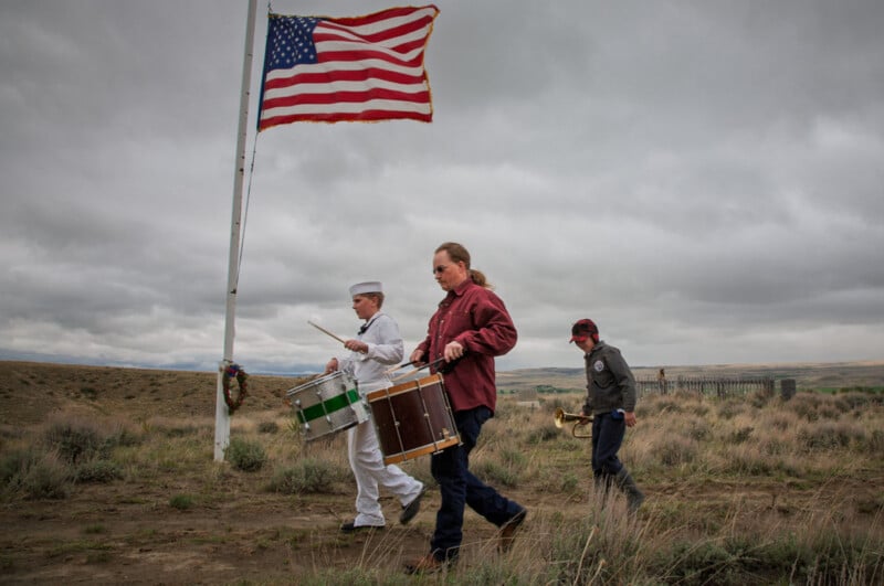 Three people, including a sailor in white and two others, walk in a grassy field carrying drums and a trumpet, with a large American flag on a pole waving in the background under a cloudy sky.