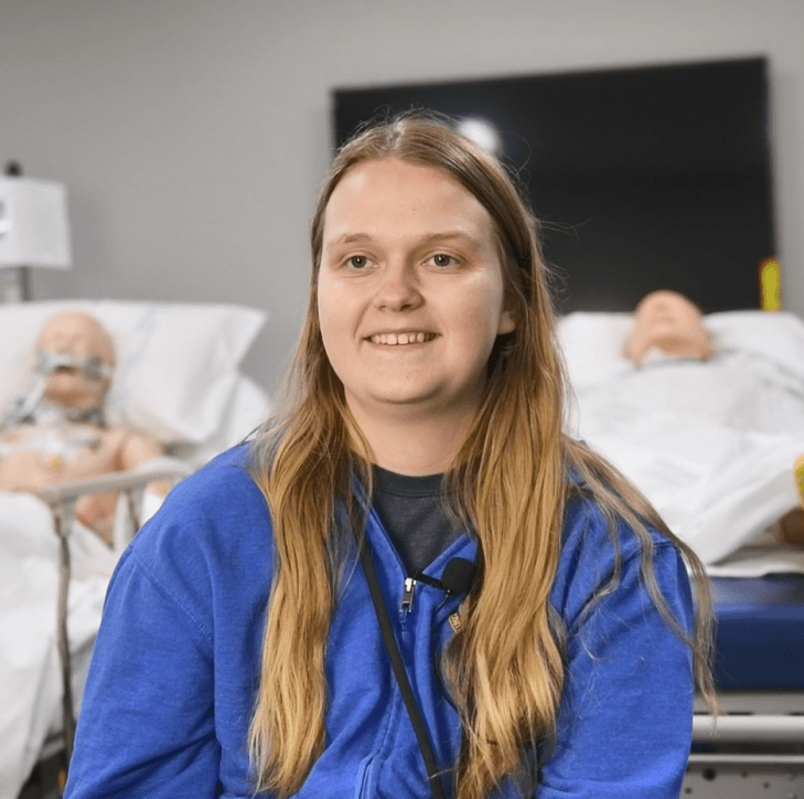 A person with long hair wearing a blue hoodie smiles in a medical setting. Behind them are two medical mannequins on hospital beds, suggesting a training environment.