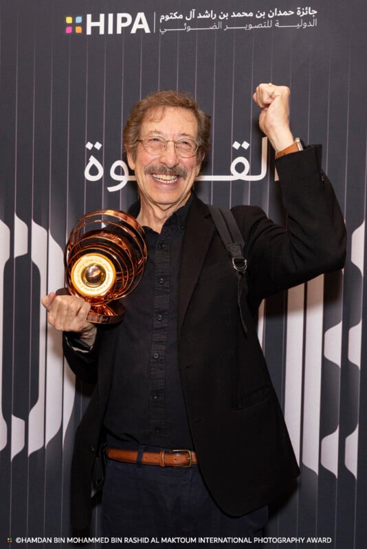 A smiling man with glasses and a mustache raises his fist in celebration while holding a shiny award. He stands in front of a backdrop with the HIPA logo and Arabic text.