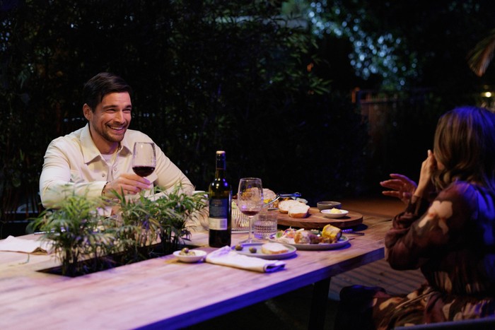 A smiling man raises a glass of red wine during an outdoor dinner at night, seated across from a woman at a table set with food, plates, and a wine bottle amid garden greenery.