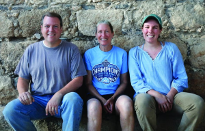 Three people sit side by side smiling at the camera, with their backs against a rock wall.