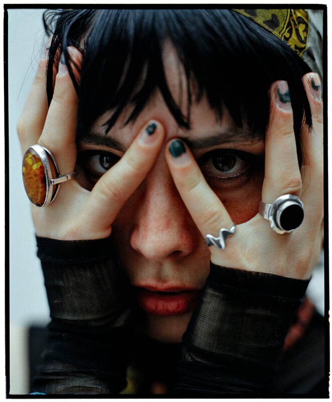 A person with black hair peers through their fingers pressed against their face, wearing statement rings and black mesh sleeves, with chipped dark nail polish and a patterned headscarf.