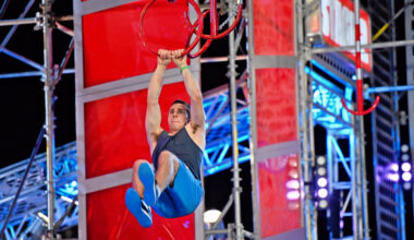 An athlete grips suspended rings mid-air on an obstacle course, wearing a dark sleeveless top and blue pants against a backdrop of dramatic red panels, metal scaffolding, and colorful stage lighting.