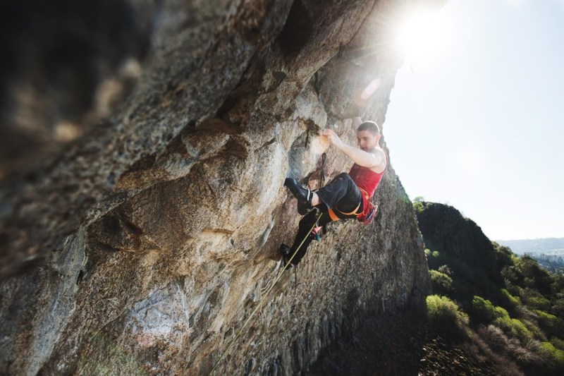 A rock climber scales a steep natural cliff face wearing safety gear and a harness, with green hillside landscape visible far below in the background.