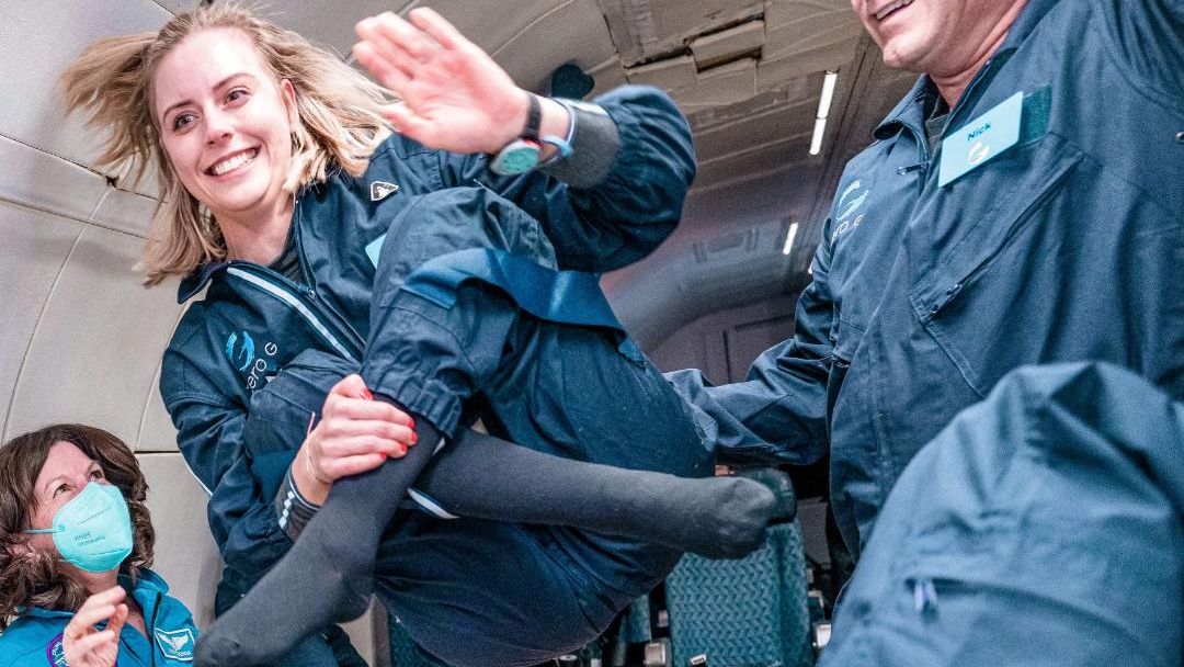 photo of a smiling and waving young woman wearing a blue jumpsuit on a plane