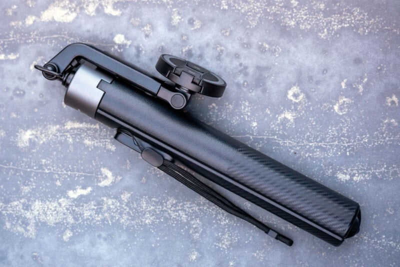 A black, compact tripod with a phone holder attachment is folded and placed on a textured gray surface with light speckles.