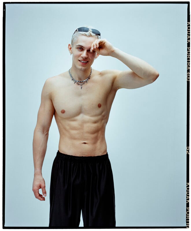 A shirtless young man with light hair stands in black shorts against a light background, smiling and touching his eyebrow with one hand. He wears a necklace and sunglasses on his head.
