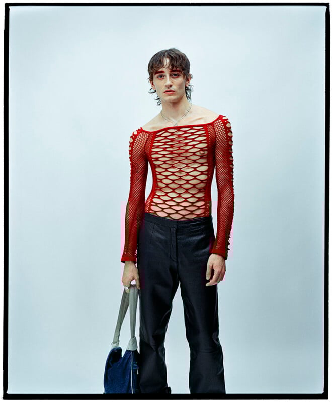 A person with shaggy hair wears a red, long-sleeve, netted top with a crisscross pattern and black pants, holding a blue and gray bag, standing against a plain light background.