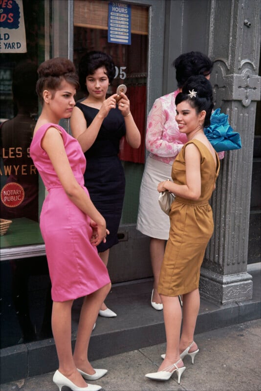 Four women stand outside a storefront. Three face the camera, wearing colorful 1960s dresses and white heels; one is in pink, another in mustard yellow, and one in black. A fourth, in white, faces away.