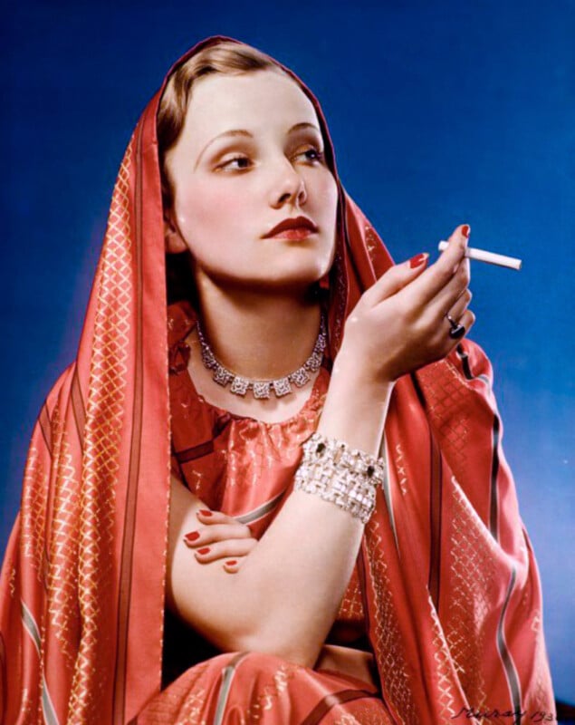 A woman wearing a red patterned sari and jewelry holds a cigarette in one hand. She has short, styled hair and red lipstick, posing against a blue background.