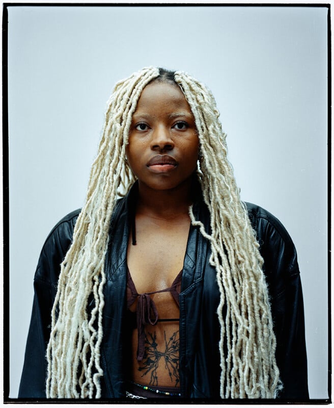 A person with long, blonde, textured braids wears a black leather jacket over a dark top, revealing a large tattoo on their chest. They look directly at the camera against a plain, light background.