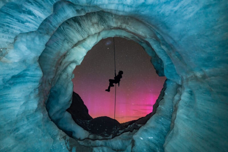 A person is rappelling inside a circular glacier cave, silhouetted against a starry sky with pink and purple aurora lights. The icy cave walls are blue and textured, framing the vibrant night sky.