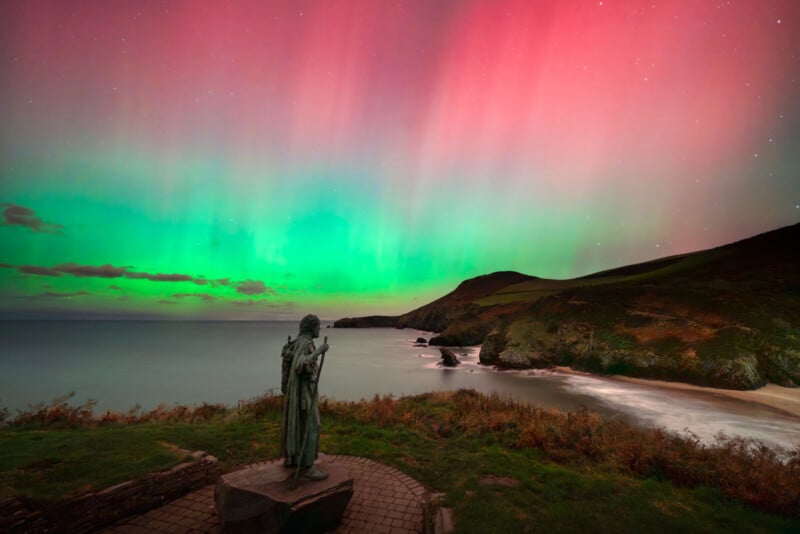 A statue overlooks a coastal landscape with cliffs and sea under a vibrant aurora, featuring green and pink lights illuminating the night sky above the shoreline.