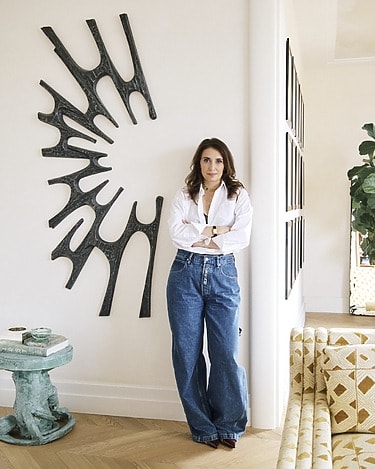 Person in a white shirt and jeans standing by abstract wall art in a stylish room with a patterned couch and plant.