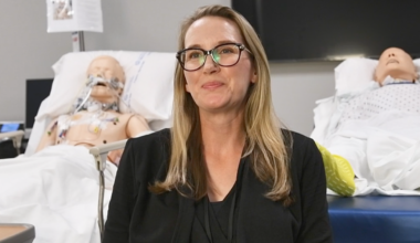 A woman with long hair and glasses smiles in a medical simulation room. Behind her are two realistic medical dummies on hospital beds, suggesting she's a trainee.