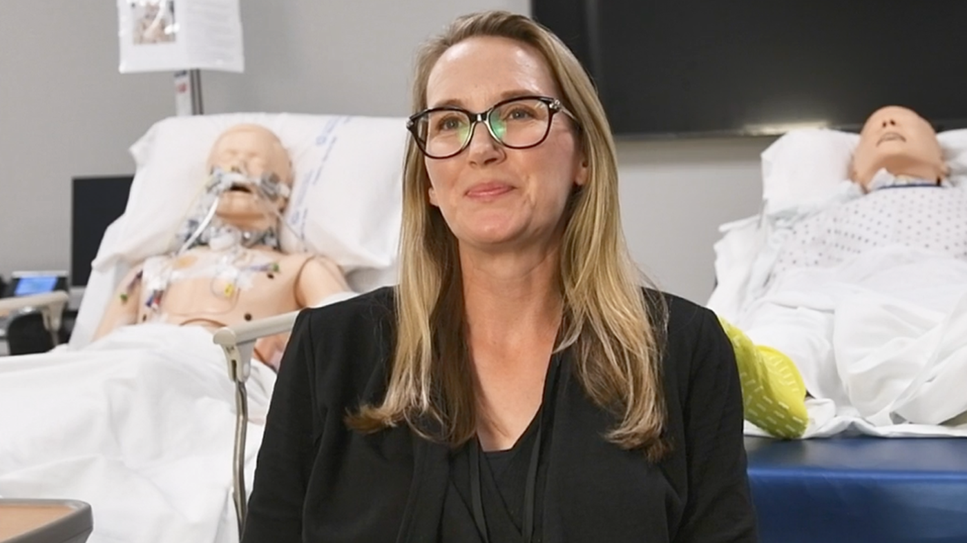 A woman with long hair and glasses smiles in a medical simulation room. Behind her are two realistic medical dummies on hospital beds, suggesting she's a trainee.