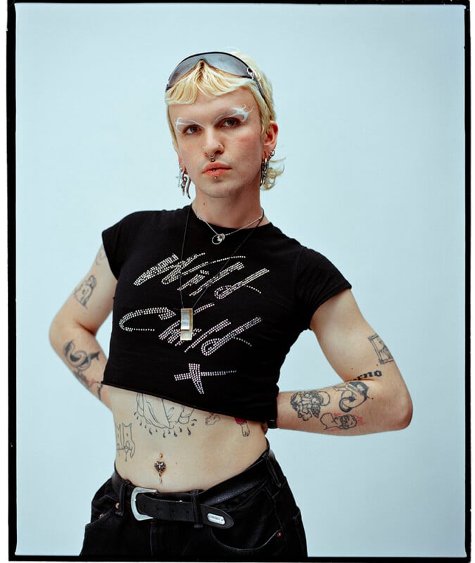 A person with bleached hair, bold makeup, and facial piercings poses confidently with hands on hips. They wear a cropped black t-shirt with silver text, dark pants, sunglasses on their head, and have visible tattoos and jewelry.