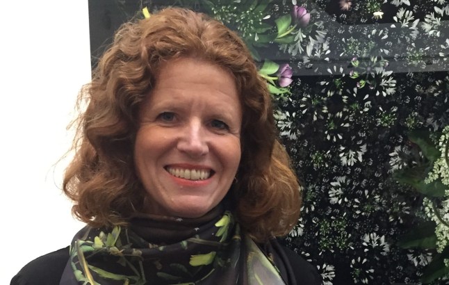 Millie Greenwood. She has auburn hair, a black scarf with a green botanical pattern and a print with flowers on behind her. She wears coral lipstick and smiles at the camera.