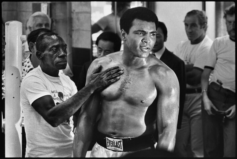 A boxer wearing Everlast shorts stands in a gym, looking determined. An older man touches his chest as several people watch in the background. The scene is intense and focused.
