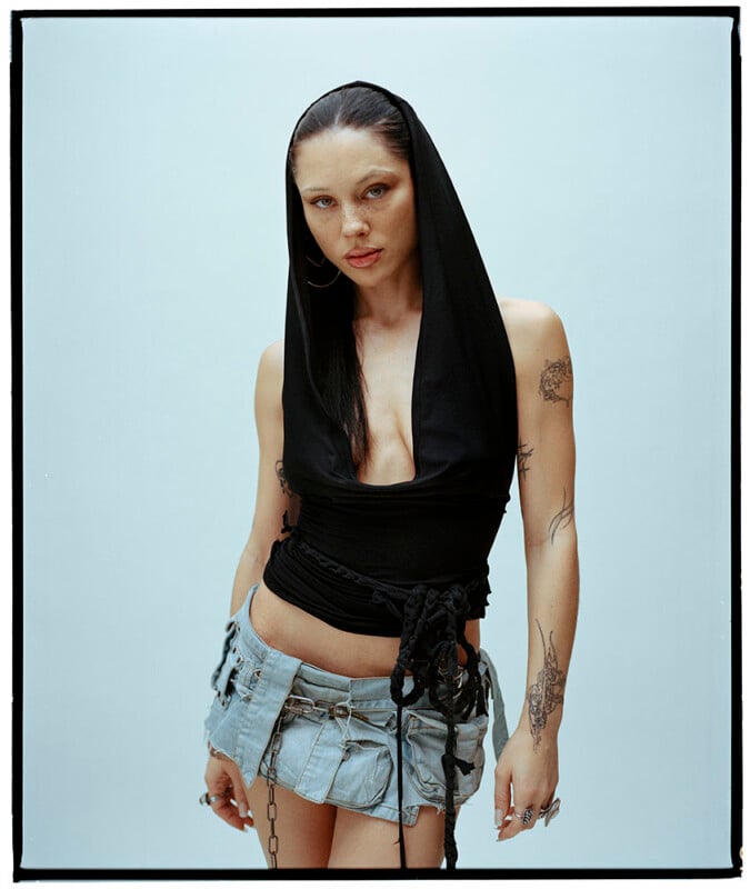 A person with long dark hair wears a sleeveless black hooded top and a short, frayed denim skirt. They have visible tattoos on their arms and stand against a plain light background, looking directly at the camera.