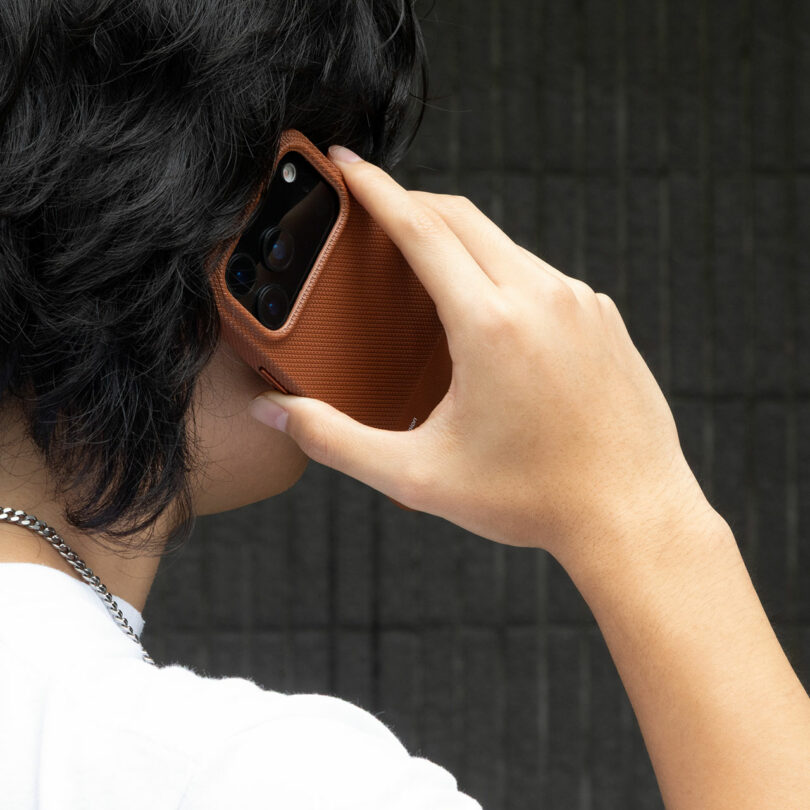 Person holding a smartphone with a brown textured case to their ear, appearing to make a call, against a dark, grid-patterned background.