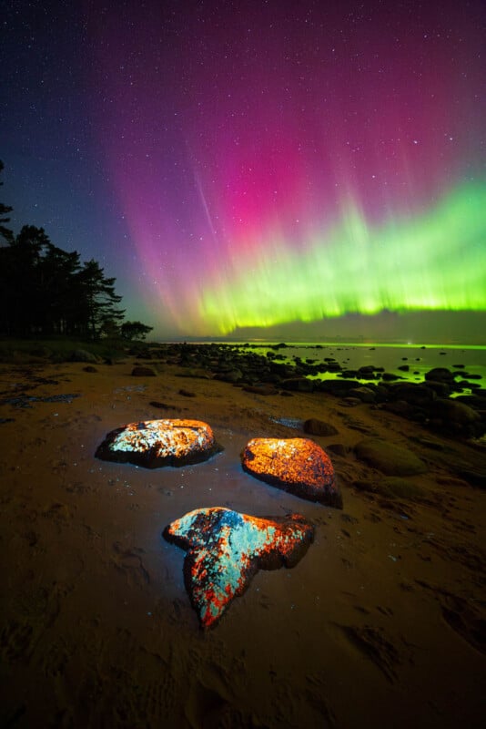Large glowing rocks on a sandy beach under a vibrant aurora borealis, with green and pink lights in the night sky and a silhouette of trees on the left.