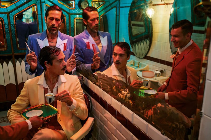 Four men in colorful suits, two in blue and two in red, stand in a retro bathroom with mirrors. One man is seated, holding a cup and saucer while the others hold grooming tools, creating a surreal, theatrical scene.