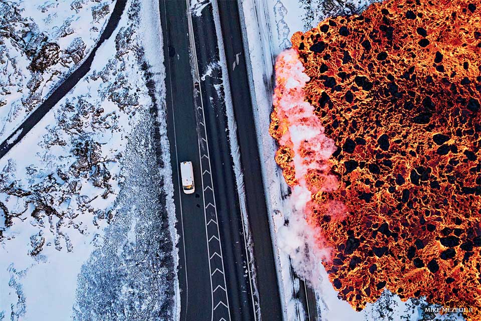 An image of a vehicle driving down the road as lava rushes towards the road.