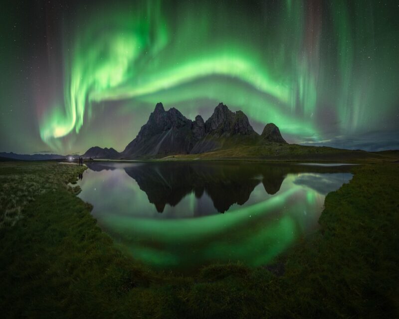 Swirling green aurora over sharp mountains with a reflection in a foreground lake.
