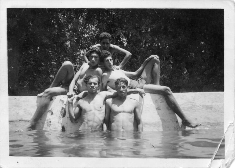 Five young men pose shirtless at the edge of a pool, three sitting on the wall and two standing in the water. Trees and foliage fill the background in this black-and-white vintage photo.