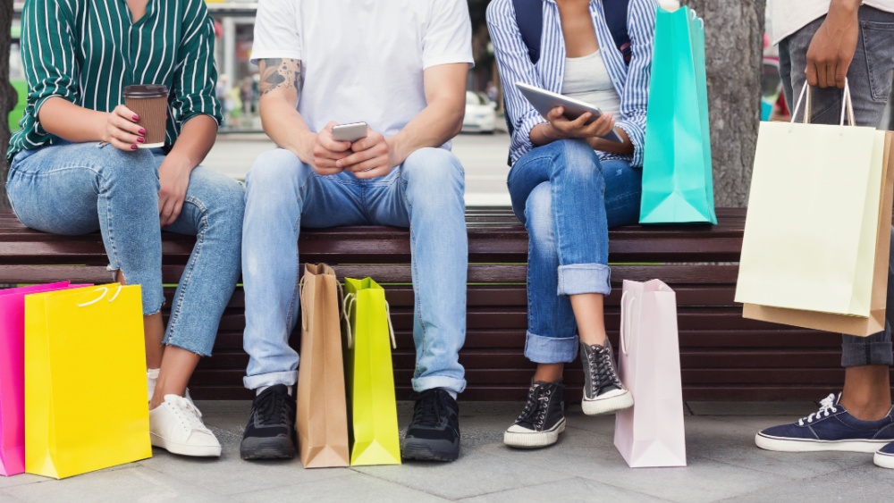 people sitting down holding phones and paper cups with paper shopping bags at their feet