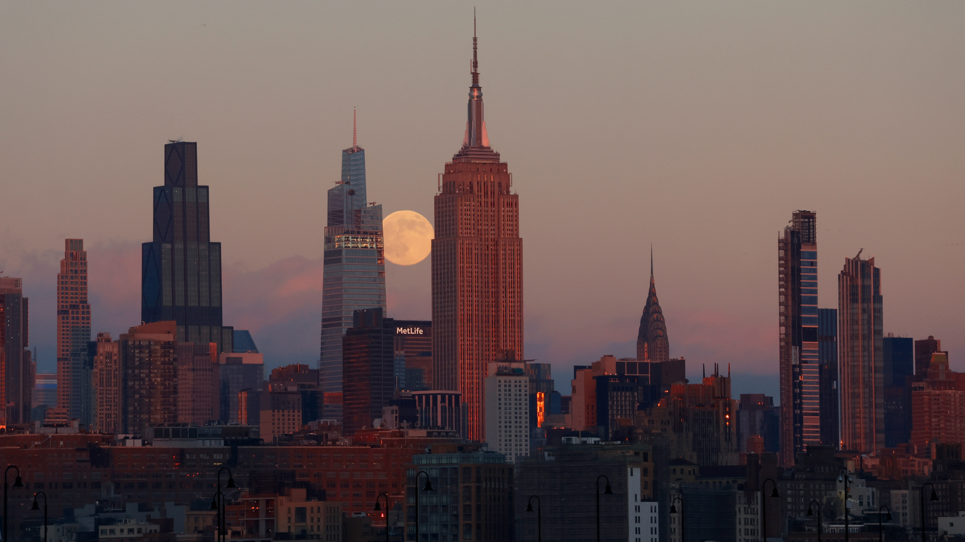 A full moon is pictured rising between the Empire State Building and another skyscraper in an evening sky, as they and other highrise buildings dominate the New York skyline.