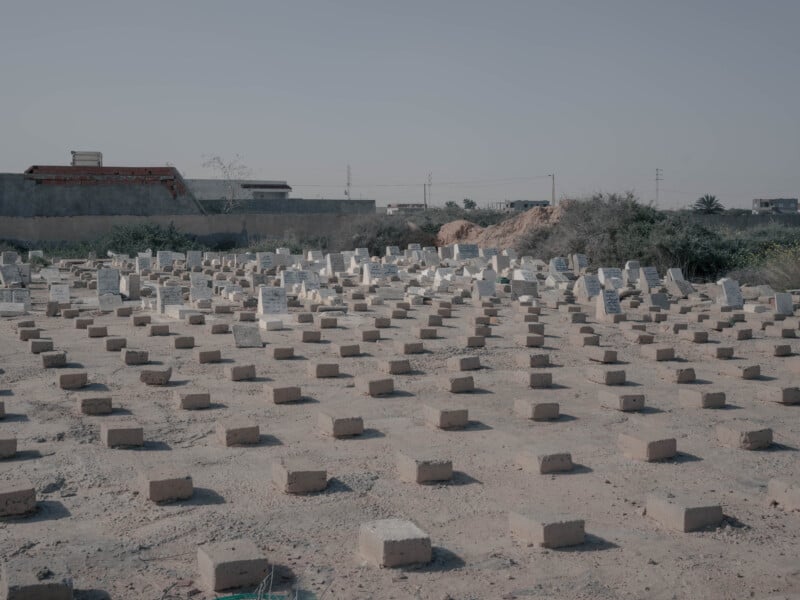 A cemetery with rows of simple, rectangular graves marked by small, light-colored headstones and bricks, set in a dry, sandy area with buildings and vegetation in the background.