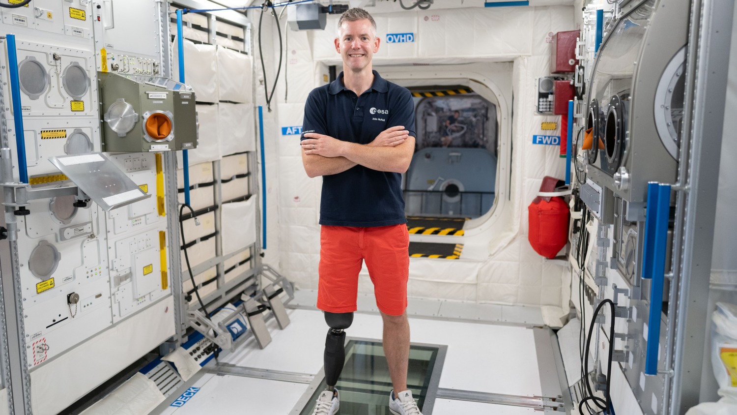 a man with a prosthetic legs stands wearing a dark blue polo shirt and orange shorts stands in a mockup International Space Station module.