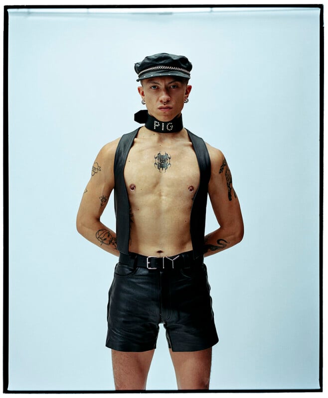 A person wearing a black leather cap, collar with "PIG" spelled in studs, sleeveless leather vest, and black leather shorts stands with hands behind their back against a plain light background. Tattoos are visible on their torso and arms.