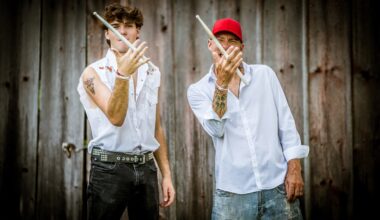 Two men in casual clothing stand in front of a wooden wall, each holding up a pair of drumsticks crossed in an X.
