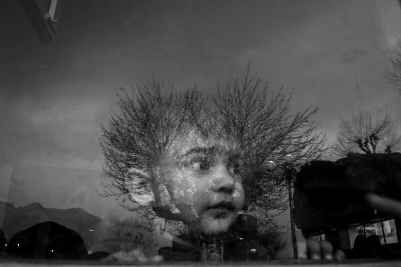 A black-and-white photo shows a child’s face reflected in a window, overlaid with leafless tree branches that appear to form a crown or hair around the child’s head, creating a surreal and dreamy effect.