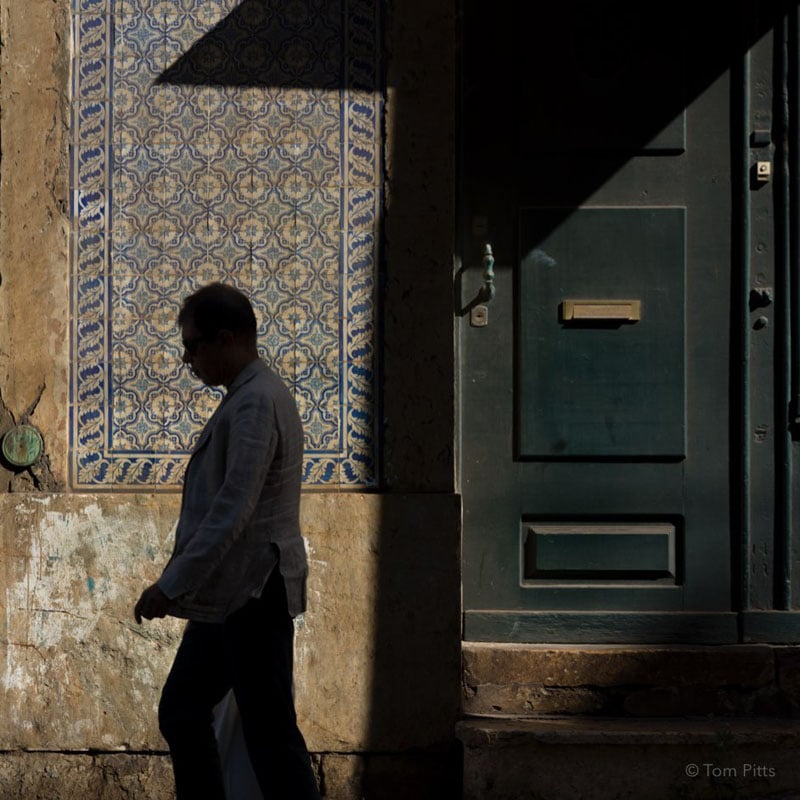 A man in silhouette walks past a sunlit tiled wall and a large dark green door, with part of the scene in deep shadow.