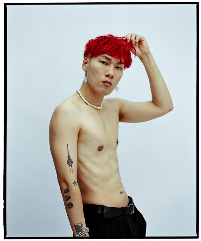 A shirtless person with bright red hair, wearing a pearl necklace and earrings, poses with one hand touching their head. They have visible tattoos on their left arm and stand against a plain light background.