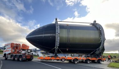 A large, metal bullet-shaped rocket is carried on a trailer behind a truck.