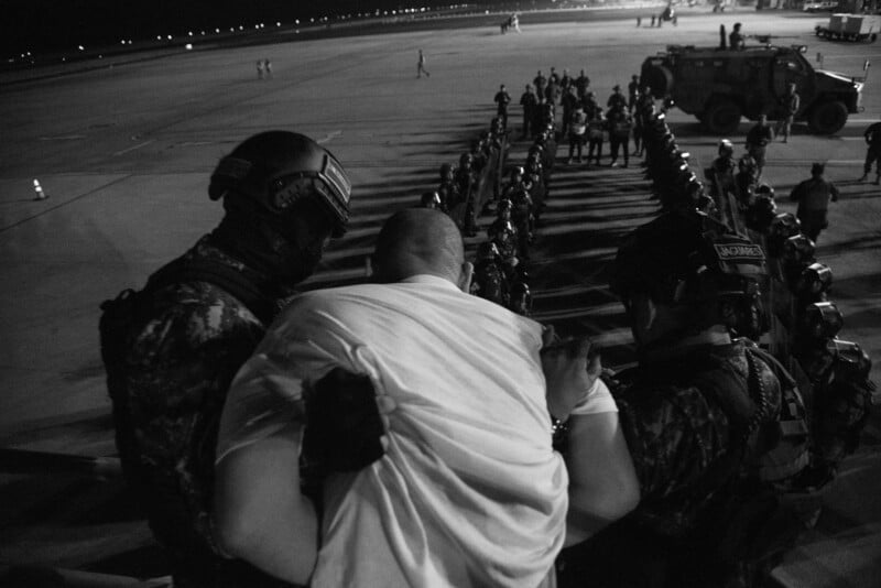 A black and white photo shows two armed officers escorting a man in a white shirt down airplane steps toward a line of uniformed personnel standing at attention on an airport tarmac at night.