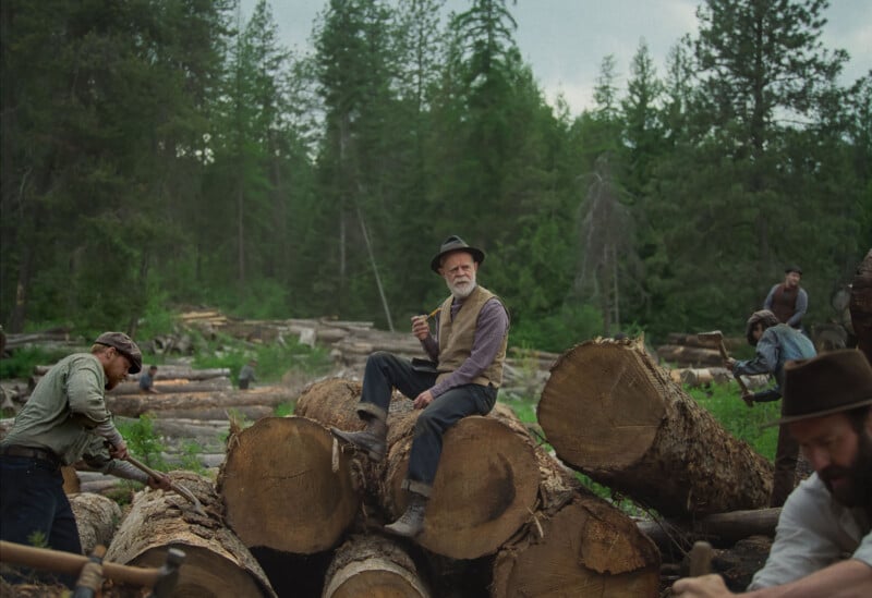 A man in old-fashioned clothes and a hat sits on large cut tree logs in a forest, holding an axe. Other men work around him, sawing and chopping logs among tall pine trees.