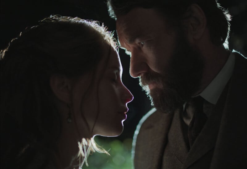 A woman and a man stand closely, facing each other in dramatic lighting. The woman has her eyes closed, and the man looks at her intently. Both are dressed in vintage-style clothing, creating an intense and emotional atmosphere.