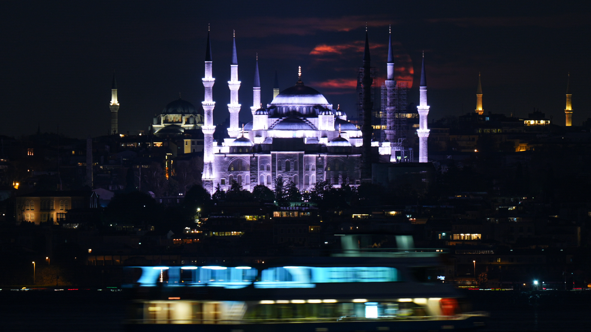 An ornate mosque is pictured lit-up at night, as a cloud-streaked red moon lurks behind it.