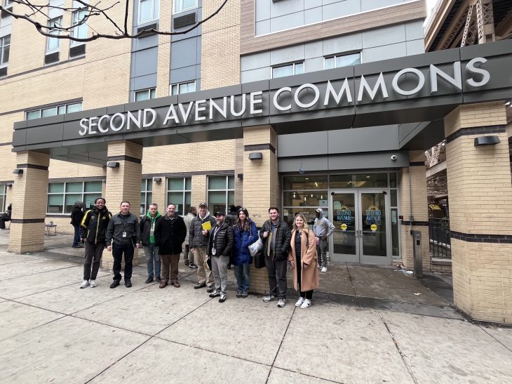 A diverse group of 11 people stand smiling in front of the "Second Avenue Commons" building. The atmosphere appears friendly and welcoming.