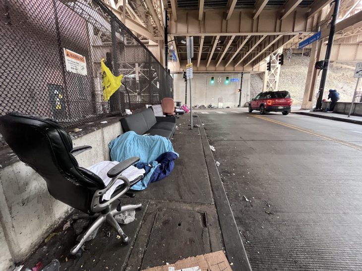 Under a bridge, discarded furniture and a blue sleeping bag suggest homelessness. A red SUV drives by on the street. The setting is gritty and somber.