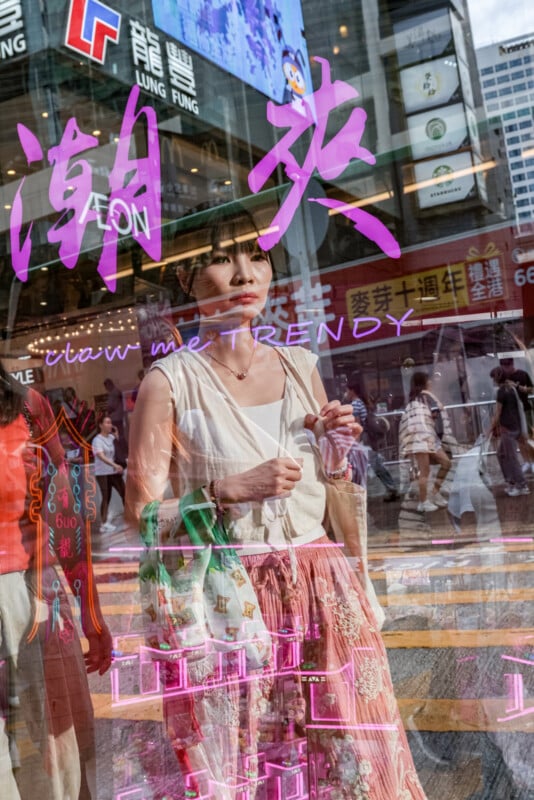 A young woman stands on a busy city street, reflected in a shop window covered with neon signs, pink and purple text, and passersby, creating a layered, vibrant urban scene.