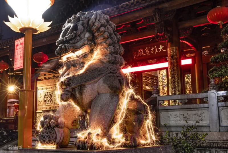 A stone Chinese guardian lion statue at night is illuminated by swirling golden light trails, with a traditional temple, red lanterns, and illuminated Chinese signs in the background.