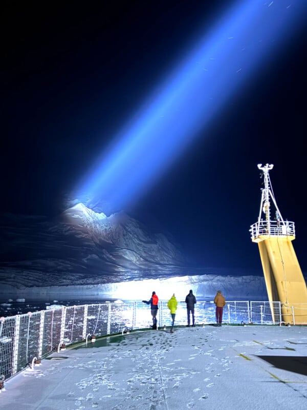 Four people stand on a ship's snowy deck at night, watching a powerful beam of light illuminate a distant, snow-covered mountain, with footprints visible on the icy surface.