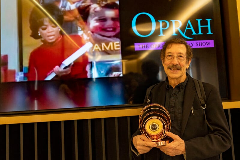 A man with gray hair and glasses smiles while holding a circular award. Behind him, a screen displays “The Oprah Winfrey Show” logo and an image of Oprah Winfrey holding a large object.